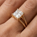 A gold ring with a square princess-cut clear gemstone in a four-prong setting, worn on a finger. The ring features a split shank band design.