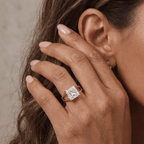 A close-up lifestyle shot of a woman's hand wearing a rose gold ring with a princess-cut diamond. The ring is shown on her ring finger, with her ear and hair visible in the background.