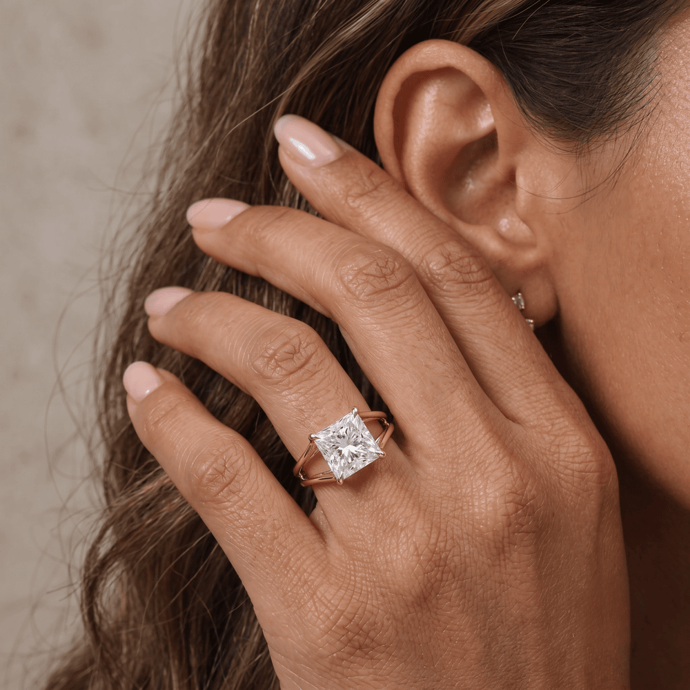 A close-up lifestyle shot of a woman's hand wearing a rose gold ring with a princess-cut diamond. The ring is shown on her ring finger, with her ear and hair visible in the background.