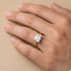 A close-up of a hand wearing a rose gold ring with a square-cut clear gemstone, set against a neutral beige background.