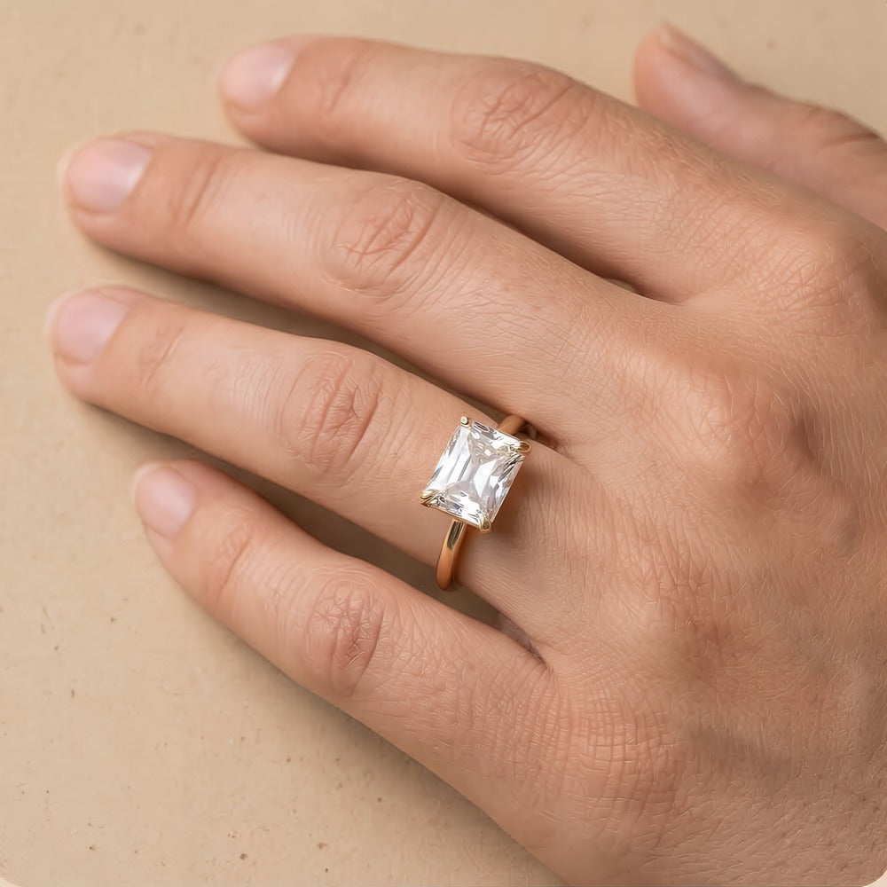 A close-up of a hand wearing a rose gold ring with a square-cut clear gemstone, set against a neutral beige background.