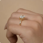 A close-up of a hand wearing a yellow gold ring with a square-cut clear gemstone in a prong setting, set against a solid beige background.