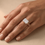 A close-up of a hand wearing a rose gold ring with a rectangular clear gemstone, set against a neutral beige background.