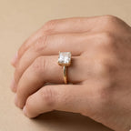 A gold ring with a square clear gemstone worn on a person's hand, set against a warm beige background.