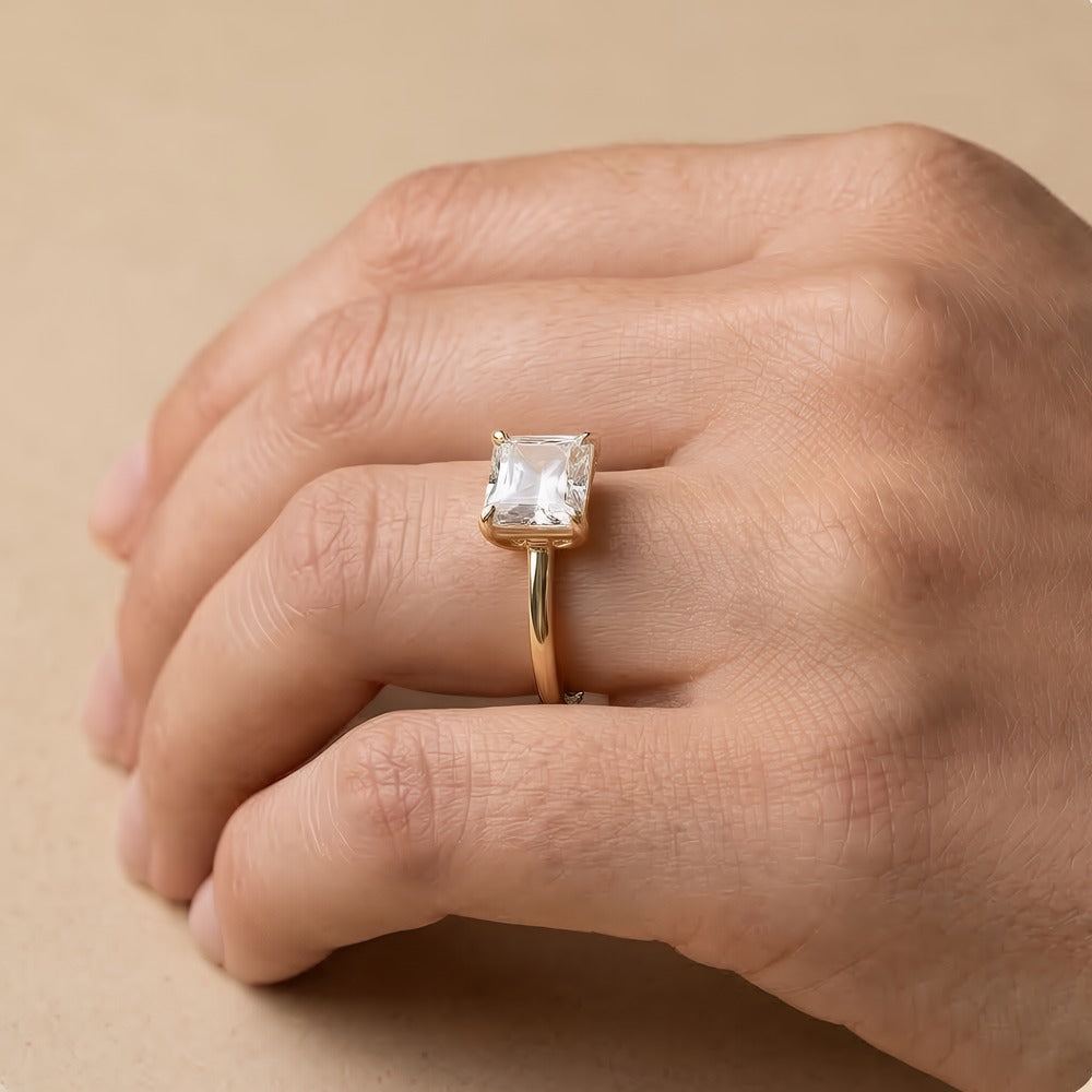 A gold ring with a square clear gemstone worn on a person's hand, set against a warm beige background.