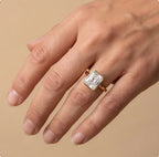 A close-up of a hand wearing a gold ring with a rectangular clear gemstone, set against a solid beige background.