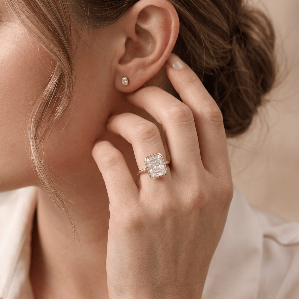 A close-up lifestyle shot of a woman wearing a rose gold ring with an emerald-cut clear gemstone and a matching small stud earring.