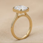 A gold ring with a square bezel holding a clear, cushion-cut gemstone, set against a neutral beige textured background.