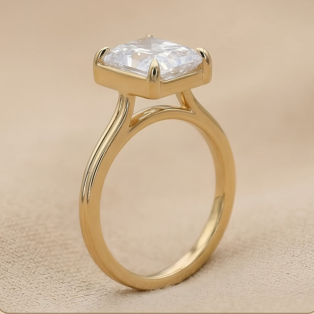 A gold ring with a square bezel holding a clear, cushion-cut gemstone, set against a neutral beige textured background.
