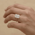 A close-up of a hand wearing a ring with a large rectangular clear gemstone in a silver-toned setting, against a warm beige background.