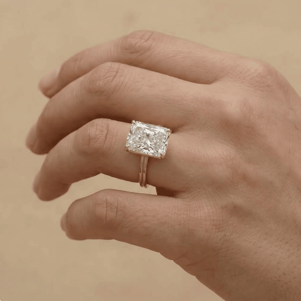 A close-up of a hand wearing a ring with a large rectangular clear gemstone in a silver-toned setting, against a warm beige background.