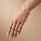 A close-up of a person's hand wearing a delicate diamond bracelet with a central round-cut diamond, set against a soft beige background.