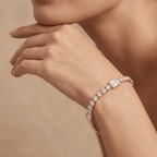 A close-up of a model's wrist wearing a yellow gold tennis bracelet adorned with clear emerald-cut and round diamonds, set against a warm beige background. 