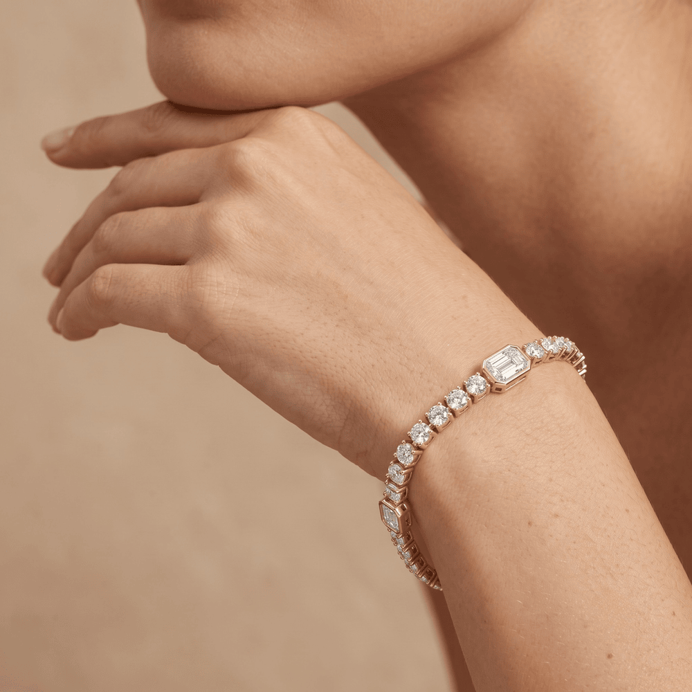 A close-up of a model's wrist wearing a yellow gold tennis bracelet adorned with clear emerald-cut and round diamonds, set against a warm beige background. 