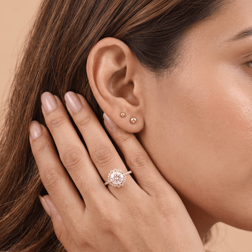 A close-up lifestyle shot of a woman's ear and hand, showcasing a rose gold halo ring with a central clear gemstone and matching rose gold stud earrings.