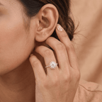 A close-up lifestyle shot of a woman wearing a silver halo engagement ring with a round-cut center stone on her finger, with her hand resting near her ear against a warm beige background.