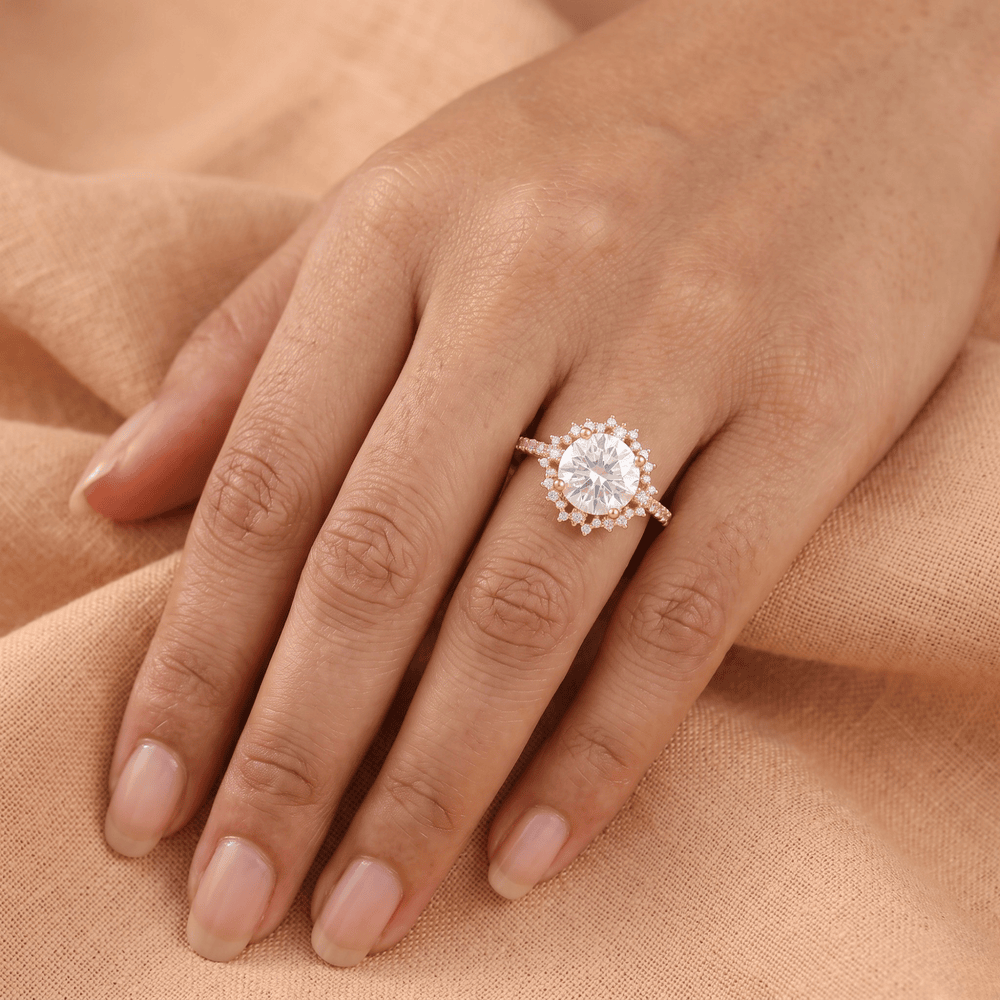 A close-up of a hand wearing a rose gold ring with a large round-cut center stone and a halo of smaller stones, resting on a soft peach fabric background.