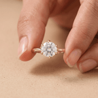 A close-up of a hand holding a rose gold solitaire engagement ring with a round brilliant-cut diamond. The ring is shown against a soft, textured beige background.