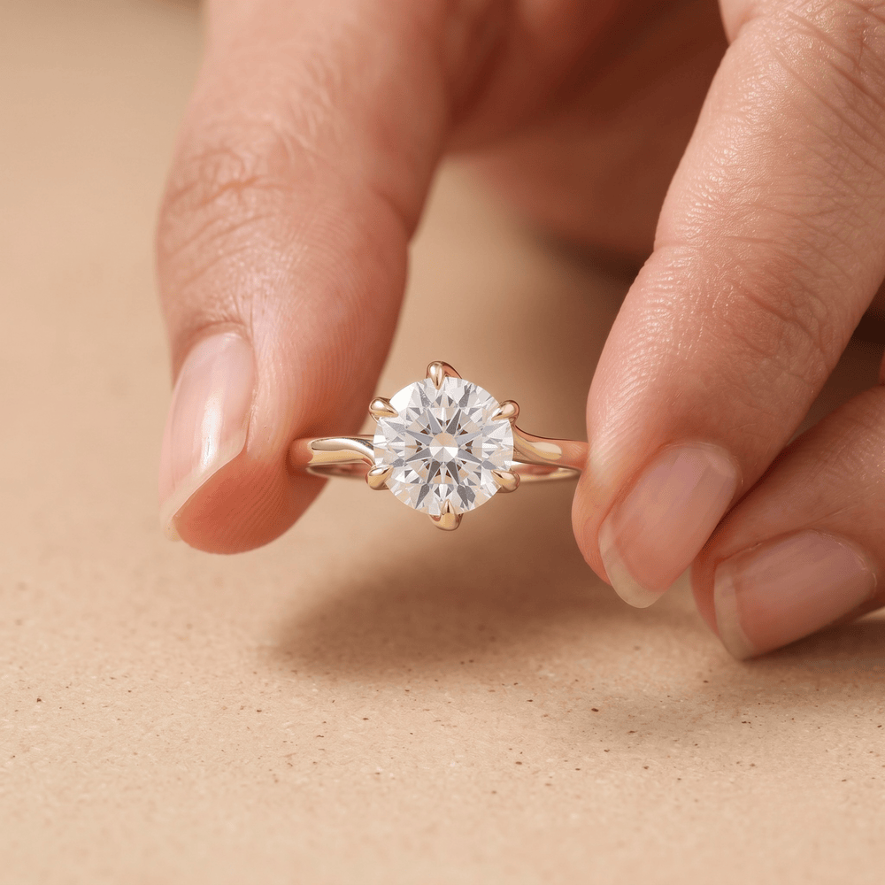 A close-up of a hand holding a rose gold solitaire engagement ring with a round brilliant-cut diamond. The ring is shown against a soft, textured beige background.