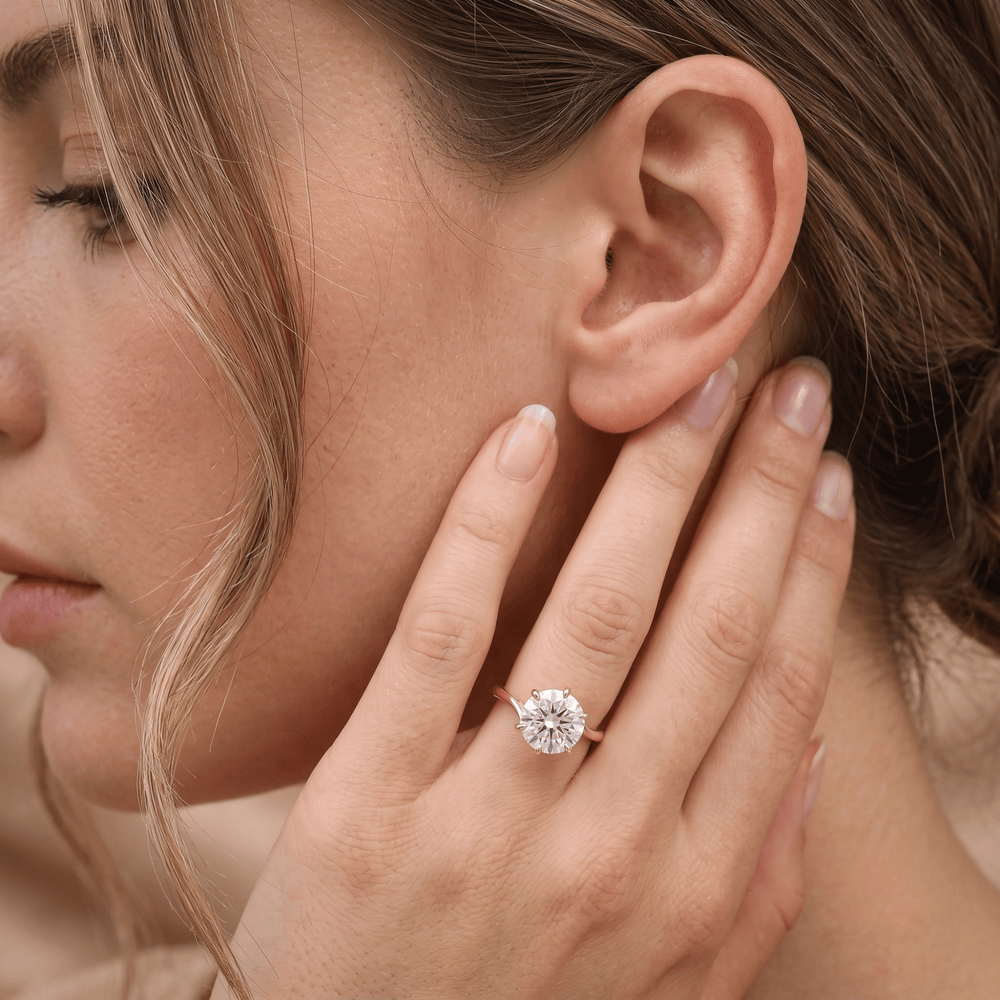 A close-up lifestyle shot of a woman wearing a classic solitaire diamond engagement ring on a rose gold band, with her hand resting near her ear.