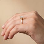 A close-up of a hand wearing a yellow gold ring with a heart-shaped clear gemstone, set against a soft beige background.