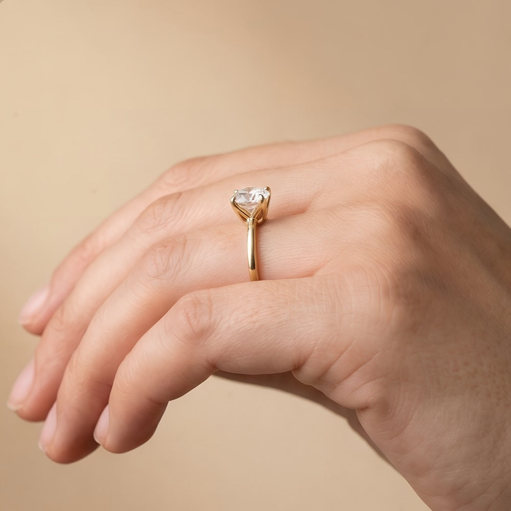 A close-up of a hand wearing a yellow gold ring with a heart-shaped clear gemstone, set against a soft beige background.