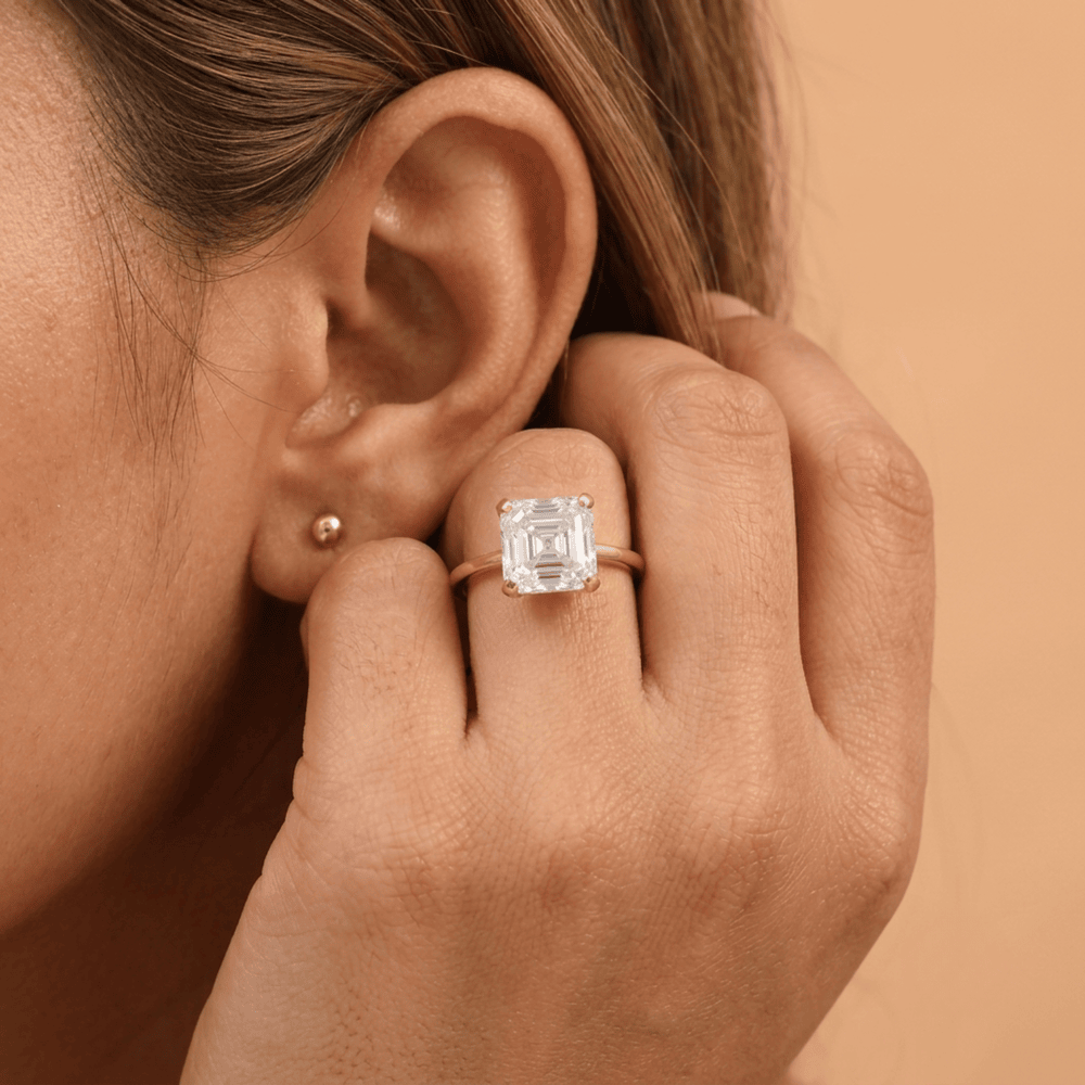 A close-up lifestyle shot of a person wearing a rose gold ring with a clear emerald-cut gemstone on their finger, alongside a small gold stud earring, set against a warm beige background.